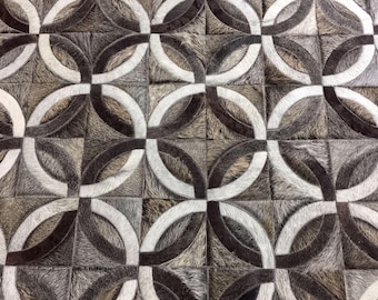 May include: A rug constructed from square patches of cowhide featuring a repeating geometric pattern of interlocking circles in shades of white, dark brown, and grey. The texture of the hide is visible throughout the surface of the rug.