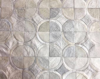 May include: A rug composed of stitched cowhide patches in shades of gray, beige, and off-white. The surface features a repeating geometric pattern of large circles intersected by straight lines, creating a textured, multi-tonal appearance.