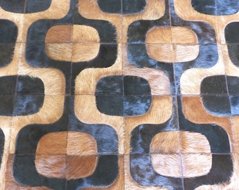 May include: A rug constructed from stitched panels of cowhide featuring a repeating geometric pattern. The design consists of interlocking rounded square shapes in alternating shades of dark brown, black, and light tan with a textured hair finish.
