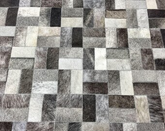 May include: A rectangular area rug constructed from individual patches of cowhide leather. The rug features a geometric pattern of alternating vertical and horizontal rectangles in varying shades of charcoal, slate gray, silver, and off-white.