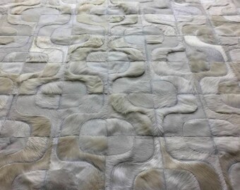 May include: A rug constructed from individual patches of light gray and beige cowhide leather. The pieces are stitched together to form a repeating pattern of interlocking curved shapes, creating a textured surface with natural hair grain.