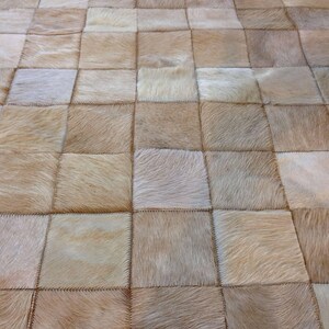 Puede incluir: Una alfombra con patrón de cuadrados construida a partir de parches individuales de piel de vaca natural. La alfombra presenta una variedad de tonos beige claro, bronceado y crema, con cada pieza cuadrada cosida con hilo oscuro visible.