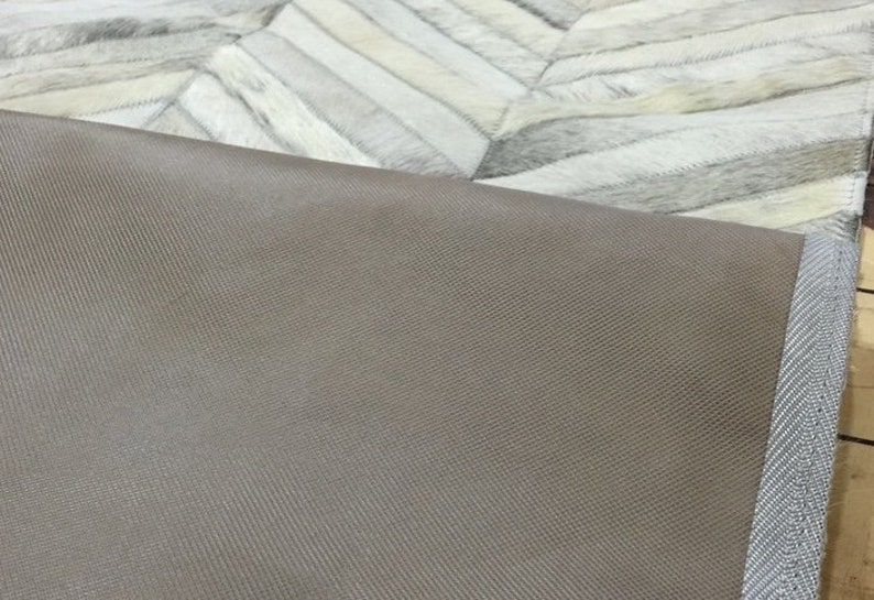 May include: Close-up of the underside of a light gray and white cowhide rug. The rug has a brown fabric backing with a gray stitched border.