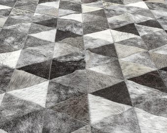 May include: A floor rug constructed from individual triangular patches of cowhide leather. The surface features a repeating geometric pattern composed of various shades of white, light gray, dark gray, and black fur, creating a textured appearance.