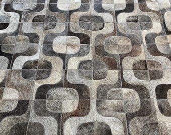 May include: A rug constructed from square patches of cowhide leather featuring a repeating geometric pattern of interlocking rounded squares in shades of dark brown, light brown, gray, and cream with a visible natural hair texture.