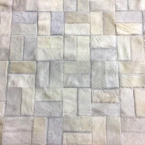 Peut inclure: Un tapis rectangulaire composé de pièces individuelles en cuir poilu. Les pièces sont disposées selon un motif répétitif en briques, présentant une palette monochrome de tons blanc cassé, crème, gris clair et argenté.