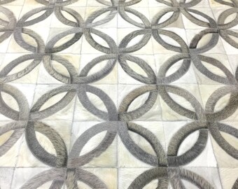 May include: A rug constructed from stitched panels of cowhide featuring a repeating geometric pattern of interlocking circles. The design alternates between shades of cream, off-white, and various tones of grey, showcasing the natural hair texture.