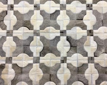 May include: A rug made of stitched cowhide patches featuring a repeating geometric pattern. The design consists of cream-colored circular and cross-shaped pieces alternating with textured gray and charcoal-colored square and rectangular hide segments.