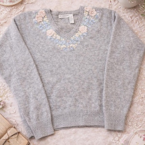May include: A pale grey V-neck jumper with floral embroidery around the neckline. The embroidery features pink, light blue, and cream-coloured flowers. The jumper is displayed on a lace surface, with a teacup and book in the background.