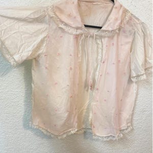 Pode incluir: Um roupão vintage rosa pálido transparente com gola e acabamentos em renda. O roupão de mangas curtas apresenta pequenos bordados florais rosa e um fecho de laço. O tecido delicado sugere uma peça de boudoir ou roupa de dormir.