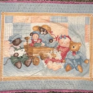 Pode incluir: Um edredom para bebé com um design de patchwork com ursinhos de peluche e outros animais de peluche. O edredom tem um fundo azul claro com uma borda rosa com babados e uma moldura dourada em torno da imagem central.