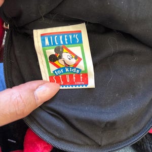 以下が含まれることがあります： 赤いリボンとジッパーが付いた黒いぬいぐるみバッグのクローズアップ。タグには「MICKEY'S for kids STUFF」とミッキーマウスのグラフィックが書かれています。バッグは柔らかい素材でできています。