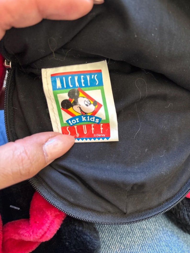 以下が含まれることがあります： 赤いリボンとジッパーが付いた黒いぬいぐるみバッグのクローズアップ。タグには「MICKEY'S for kids STUFF」とミッキーマウスのグラフィックが書かれています。バッグは柔らかい素材でできています。