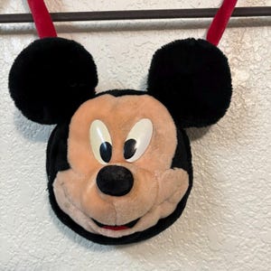 以下が含まれることがあります： 黒い耳と赤いストラップが付いた、ミッキーマウスの顔の形をしたぬいぐるみバッグ。顔はタン色で、白い目、黒い鼻、赤い笑顔があります。バッグは丸く、ヴィンテージアイテムのようです。