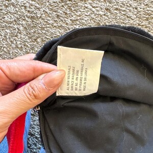 以下が含まれることがあります： ジッパー付きの黒いハンドバッグ。タグには「ALL NEW MATERIALS SURFACE WASHABLE REF. NO.: RN 84990 BY PYRAMID HANDBAGS, INC. MADE IN SRI LANKA」と記載されています。バッグには赤と青のストラップが付いています。