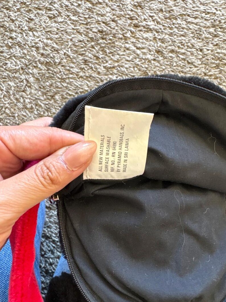 以下が含まれることがあります： ジッパー付きの黒いハンドバッグ。タグには「ALL NEW MATERIALS SURFACE WASHABLE REF. NO.: RN 84990 BY PYRAMID HANDBAGS, INC. MADE IN SRI LANKA」と記載されています。バッグには赤と青のストラップが付いています。