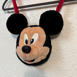 以下が含まれることがあります： 黒い耳と赤いストラップが付いた、ミッキーマウスの形をしたぬいぐるみバッグ。バッグは、ベージュの顔、黒い鼻、白い目を特徴としています。テクスチャ加工された白い壁を背景に、黒いハンガーに吊るされています。