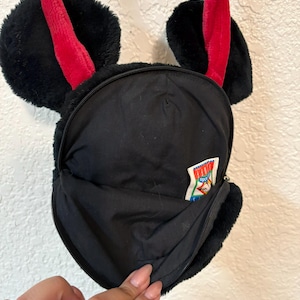 以下が含まれることがあります： 赤いストラップと耳の形をしたアクセントが付いた、黒いぬいぐるみクロスボディバッグ。バッグにはジッパー付きのメインコンパートメントと小さなジッパー付きポケットがあります。テキストの入った小さなタグが側面に表示されます。