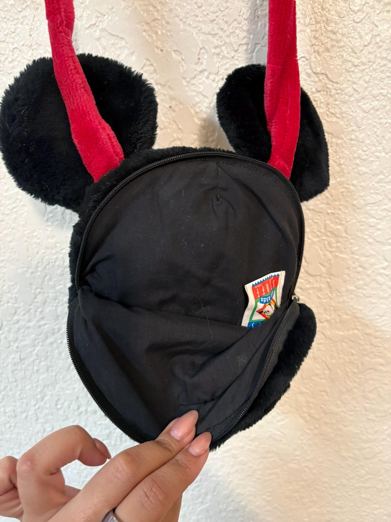 以下が含まれることがあります： 赤いストラップと耳の形をしたアクセントが付いた、黒いぬいぐるみクロスボディバッグ。バッグにはジッパー付きのメインコンパートメントと小さなジッパー付きポケットがあります。テキストの入った小さなタグが側面に表示されます。