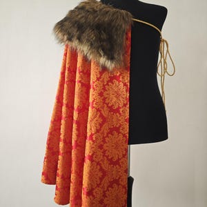 May include: A red and orange patterned cape with a faux fur collar is displayed on a black mannequin. The cape has a floral design and is secured with a gold-coloured cord. The faux fur collar is brown.