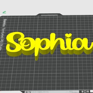 May include: A 3D-printed yellow name, "Sophia", with a heart above the "i". The name is displayed on a black grid surface, with the text "Bambu Textured PEI Plate" visible in the corner. The name is in a cursive font.