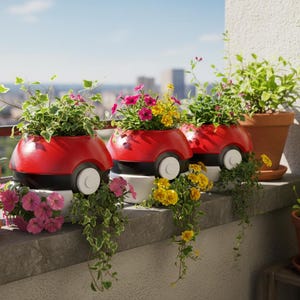 Può includere: Tre fioriere rosse e nere a forma di Poké Ball, piene di fiori colorati e verde. Le fioriere hanno ruote bianche e sono disposte su un davanzale con altri vasi di terracotta. Lo sfondo mostra un paesaggio urbano sotto un cielo azzurro.