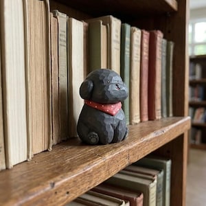 May include: A small, dark grey carved wooden figurine of a seated dog with a textured surface. The dog features a red bandana with white polka dots around its neck, small black eyes, and a tiny nose. The figure is positioned on a wooden shelf.