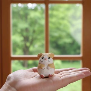 May include: A small, hand-carved wooden hamster figurine. The hamster is painted with tan, white, and pink details, and is held in the palm of a hand. The background features a window with a view of green foliage.