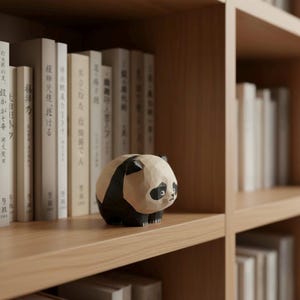 Puede incluir: Una figura de panda de madera tallada, pintada en blanco y negro, se exhibe en una estantería de madera. El panda está frente a una fila de libros con texto japonés. La estantería forma parte de una librería.