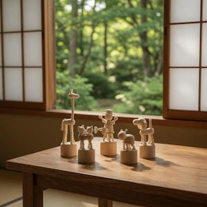 May include: A collection of wooden animal figurines, including a giraffe, bears, a person, a dog, and a beetle, are displayed on a wooden table. The figurines are light-colored and stand on round bases. The background features a window with a view of lush greenery.