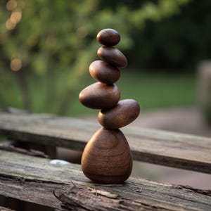 May include: A wooden sculpture of stacked, oval-shaped pieces, creating a balanced tower. The sculpture is made of dark brown wood, with a smooth, polished finish. The background is blurred, showing a green, natural setting.