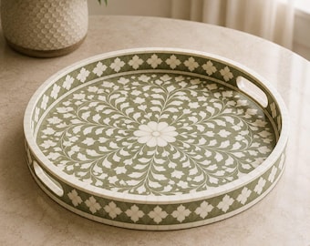 Peut inclure: Un plateau de service rond présentant un fond vert sauge avec un motif floral blanc complexe rayonnant depuis le centre. Le plateau possède des bords surélevés avec un motif quadrilobé blanc répétitif et deux poignées latérales.