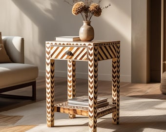 Peut inclure: Une table d'appoint carrée avec un motif en chevrons marron et blanc sur ses quatre pieds et la façade du tiroir. La table comprend un tiroir avec un bouton sombre, une étagère inférieure avec des livres et un plateau avec un vase et un livre.