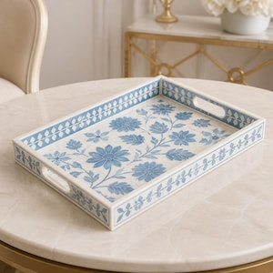Puede incluir: Una bandeja rectangular con base blanca y un patrón floral azul de flores y hojas. La bandeja tiene lados elevados con asas integradas y un diseño de borde geométrico azul y blanco que se repite a lo largo de los bordes exteriores.