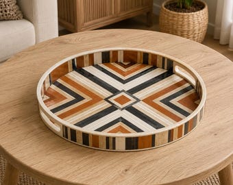 Peut inclure: Un plateau de service rond en bois présentant un motif géométrique de bandes incrustées dans des tons crème, gris anthracite et brun chaud. Le plateau possède un rebord surélevé avec deux poignées découpées intégrées et une finition lisse.