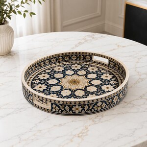 May include: A round decorative tray featuring an intricate floral pattern in shades of navy blue, cream, and gold. The tray has a raised edge with two integrated cutout handles and a detailed mosaic-style design covering the entire surface.