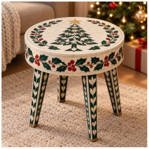 May include: A round stool with a cream-colored surface featuring an inlaid green Christmas tree, a gold star, and a wreath of green holly leaves with red berries. The sides and four tapered legs are decorated with matching holly patterns.