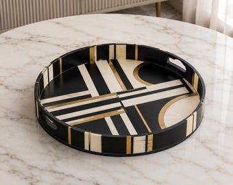 Peut inclure: Un plateau de service rond présentant un motif géométrique composé de rayures et de formes semi-circulaires noires, crème et dorées. Le plateau possède un bord surélevé avec des poignées intégrées et une finition lisse.