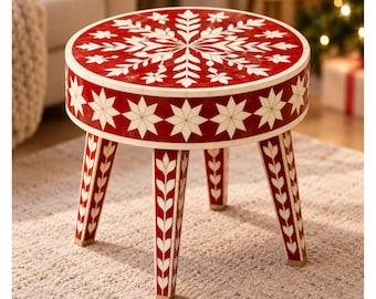 Peut inclure: Un tabouret rond présentant une surface rouge vive avec un motif de flocon de neige blanc sur le dessus. Le côté du tabouret et ses quatre pieds fuselés sont décorés de motifs d'étoiles et de feuilles blancs. La base de chaque pied est dorée.
