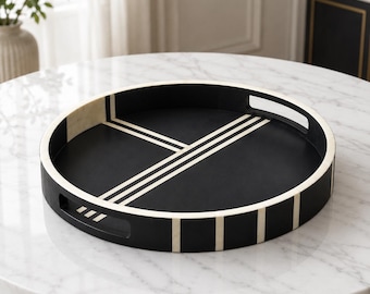 Peut inclure: Un plateau de service rond noir avec un motif géométrique incrusté blanc contrastant. Le plateau présente une finition mate lisse, un bord extérieur surélevé avec des rayures blanches verticales et deux poignées découpées intégrées.