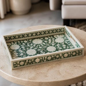 Puede incluir: Una bandeja rectangular con un fondo verde intenso y un intrincado patrón floral blanco incrustado. La bandeja tiene lados elevados con asas integradas y un borde floral blanco continuo a lo largo de los bordes exteriores.