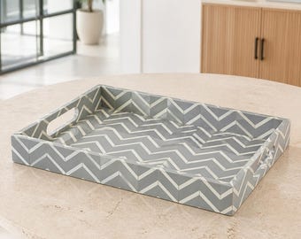 Peut inclure: Un plateau de service rectangulaire présentant un motif chevron gris et blanc répétitif sur ses surfaces intérieures et extérieures. Le plateau est doté de poignées découpées intégrées sur deux côtés opposés et d'un rebord bas.