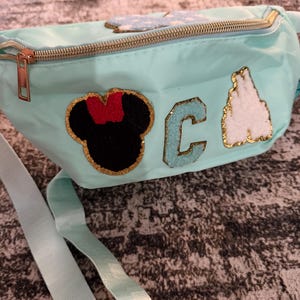 Op de afbeelding: Een lichtblauwe heuptas met een gouden rits en riem. De tas heeft patches van een zwarte Minnie Mouse-kop met een rode strik, de letter "C" en een wit kasteel, allemaal met gouden rand. De riem is lichtblauw.