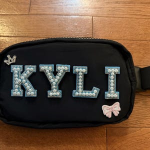 Op de afbeelding: Zwarte heuptas met de naam "KYLI" in met parels bezette letters. Een zilveren kroon en een roze strik zijn ook bevestigd. De tas heeft een zwarte riem en een ritssluiting.