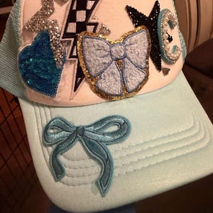 Puede incluir: Gorra de camionero azul claro con un panel frontal blanco, adornada con un corazón de lentejuelas, un rayo, un lazo y una estrella. La gorra también presenta un lazo en la visera y la letra "C".