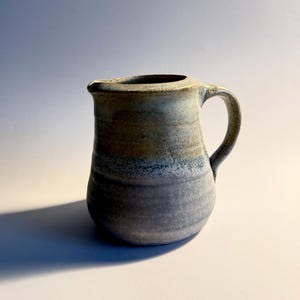 May include: A handmade ceramic pitcher with a handle and a spout. The pitcher has a rounded body and is glazed in shades of gray, blue, and tan. The pitcher is likely used for serving beverages or as a decorative item.