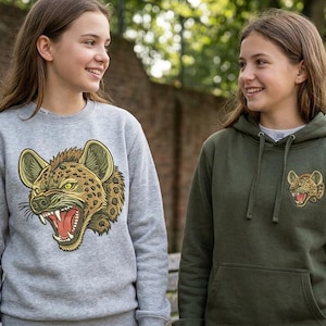 Puede incluir: Una sudadera gris claro de cuello redondo con una gran ilustración detallada de una cabeza de hiena gruñendo en el pecho, junto a una sudadera con capucha verde oliva con un gráfico de hiena más pequeño y a juego en el lado izquierdo del pecho.