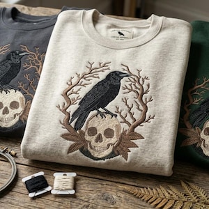Puede incluir: Tres sudaderas en carbón, crema y verde bosque, cada una bordada con un cuervo negro posado sobre una calavera, rodeado de ramas. Las sudaderas están dobladas sobre una superficie de madera, con suministros de bordado en primer plano.