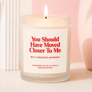 May include: A frosted glass candle with a lit wick and a white label featuring red text that reads: You Should Have Moved Closer To Me, BUT CONGRATS ANYWAYS, HANDPOURED | NET WT. 7oz | VANILLA, SipTheMomentBoutique. The candle emits a warm, steady flame.