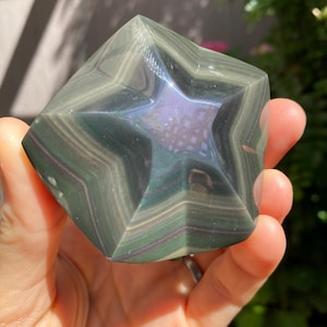 May include: A polished star-shaped stone carving featuring layered bands of dark green, black, and subtle purple hues. The surface is smooth and reflective, showcasing a distinct five-pointed star pattern formed by the natural mineral banding.